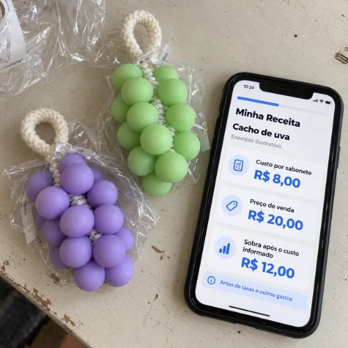 Celular com a ferramenta Minha Receita aberta, mostrando custo por sabonete, preço de venda e sobra no exemplo do cacho de uva, ao lado de dois sabonetes de cacho de uva embalados